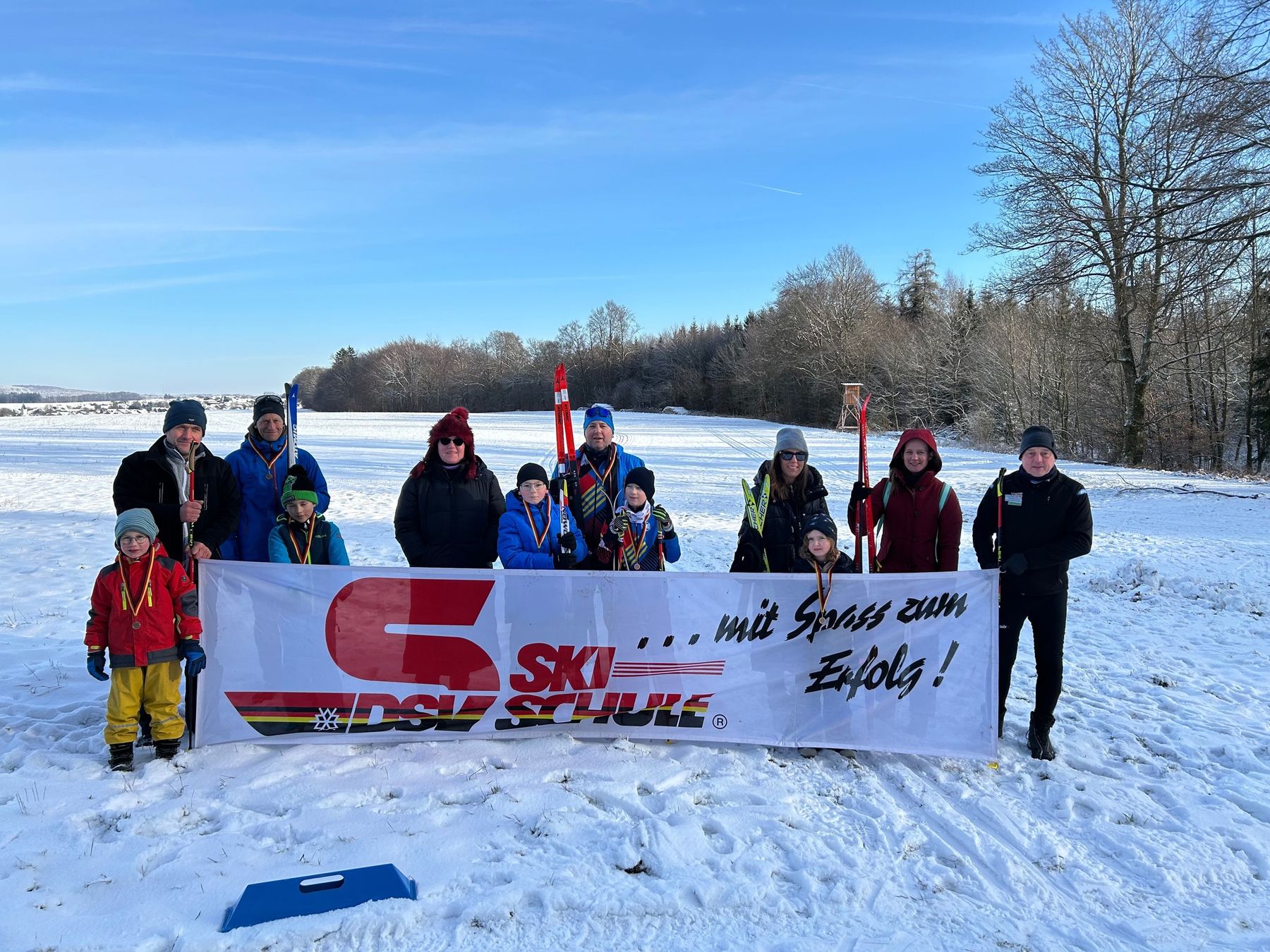 A group of adults and children dressed in winter clothing stand on snowy ground, holding skis and poles, behind a banner that reads DSV SKI SCHULE ... mit Spaß zum Erfolg! Trees and a clear sky are in the background.